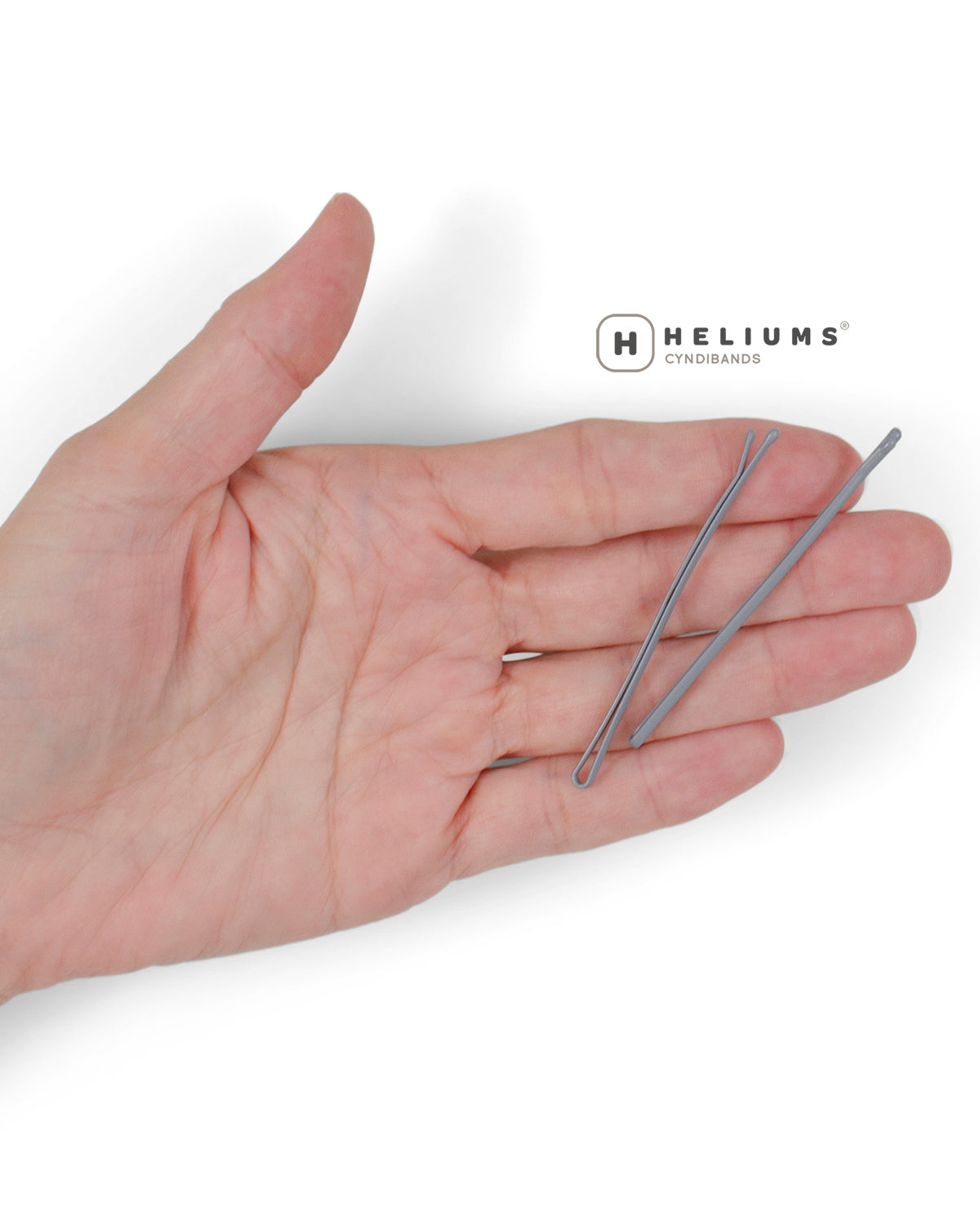 A hand holding two thin metallic bobby pins, with the Heliums brand logo printed above on a clean white background.